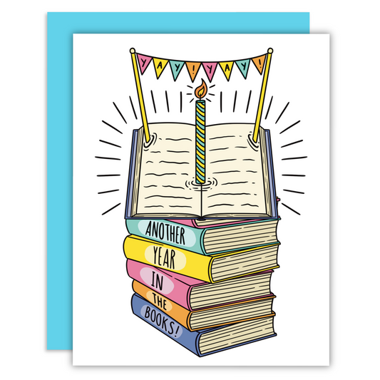 A stack of books that say another year in the books with an open book on top and a candle on a greeting card