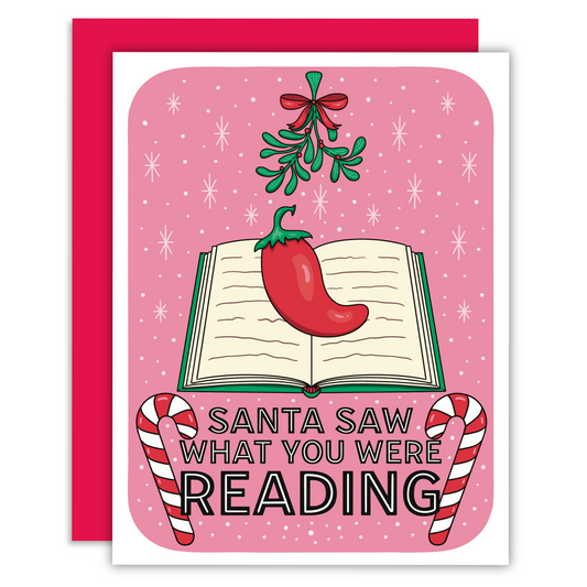 A bookish christmas greeting card with an open book and a chili pepper and a mistletoe above with snowflakes on a pink background and text that says ‘santa saw what you were reading.’