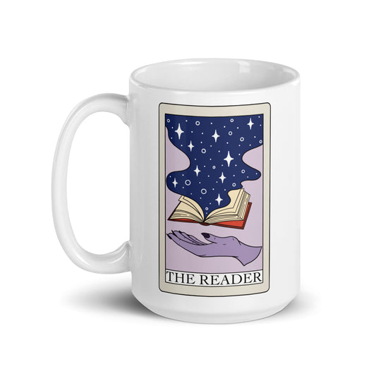 A purple hand with navy colored nails underneath a book with magic and stars spilling out of it on a tarot card. The tarot card is labeled ‘the reader’ and the design is printed on a white ceramic mug.