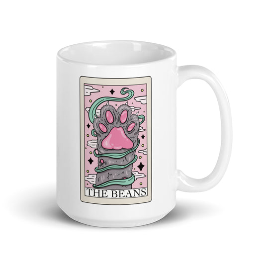  A gray paw held up with pink toe beans on a tarot card that is printed on a white ceramic mug with the text ‘the beans.’